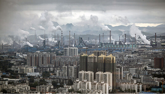 Dense industrial city with factories and smokestacks emitting thick white smoke above residential and commercial buildings under cloudy sky.