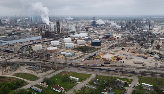 Large oil refinery with storage tanks, distillation towers, smoke stacks, and extensive piping networks under cloudy sky.