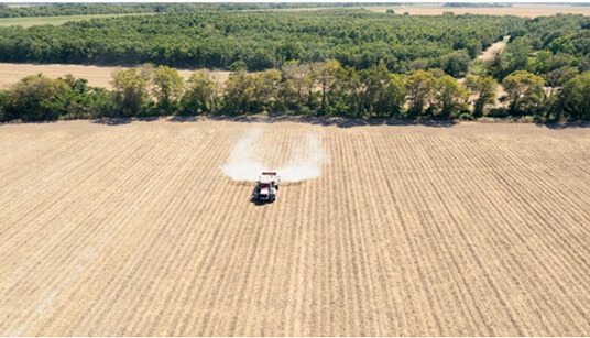 Aerial view of tractor spraying mist across evenly spaced rows in large cultivated field with tree line in background.