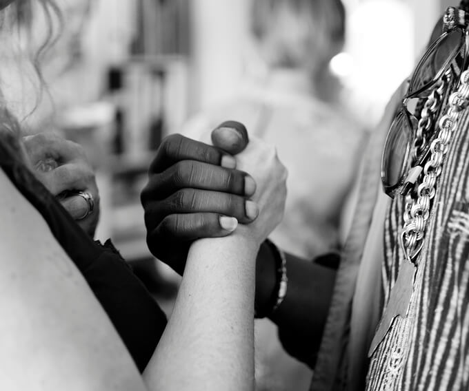 Black and white photograph of two clasped hands, one with darker skin tone and one with lighter skin tone, symbolizing unity, solidarity, and mutual respect in an indoor setting with softly blurred background.